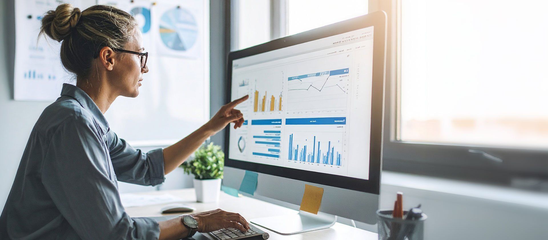 A woman sits at a desk in an office, pointing at a computer monitor displaying financial data, with pie charts mounted on the wall behind her.